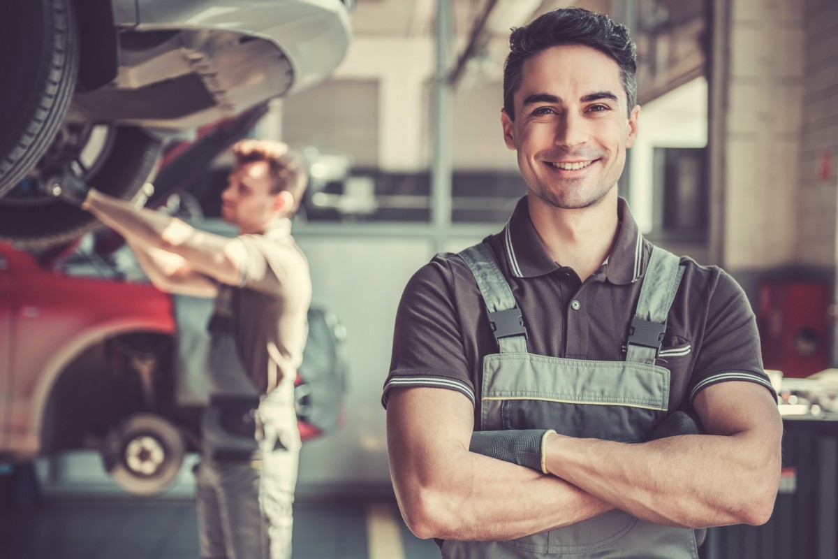 mechanic smiling in front of car on lift and another mechanic working on it 